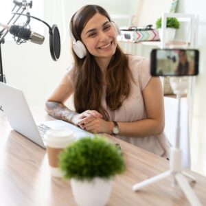 Lächelnde Podcasterin mit Kopfhörern spricht vor Mikrofon in Kamera, sitzt am Schreibtisch mit Laptop, Kaffee und Zimmerpflanze – Content Creation im Home-Studio.